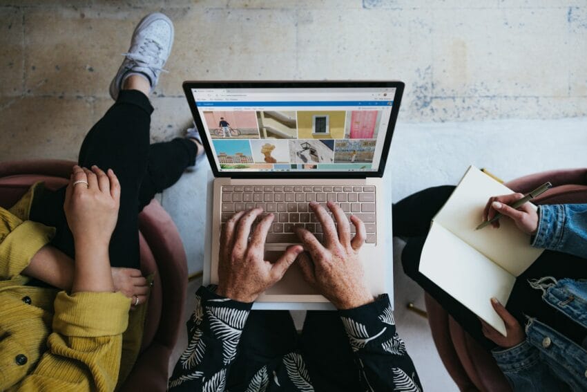 Three people gathered around a laptop, one typing while another writes in a notebook